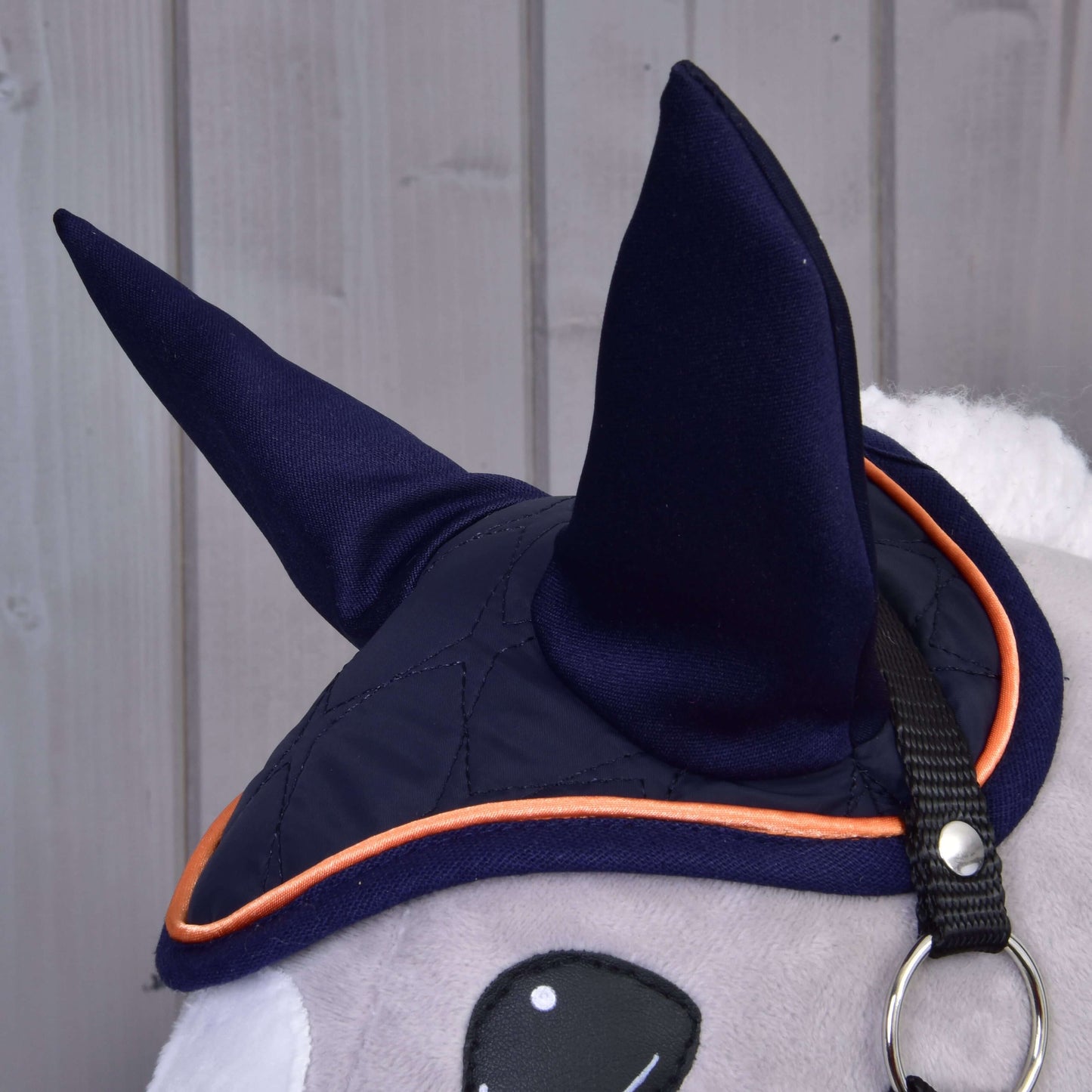 Detail Set Winner od Hobby Horse LarDen zobrazuje plyšové zvíře v tmavě modré čabrace se špičatými ušima a oranžovým lemem na pozadí světle šedých dřevěných panelů pro půvabný vzhled koňské dekorace.
