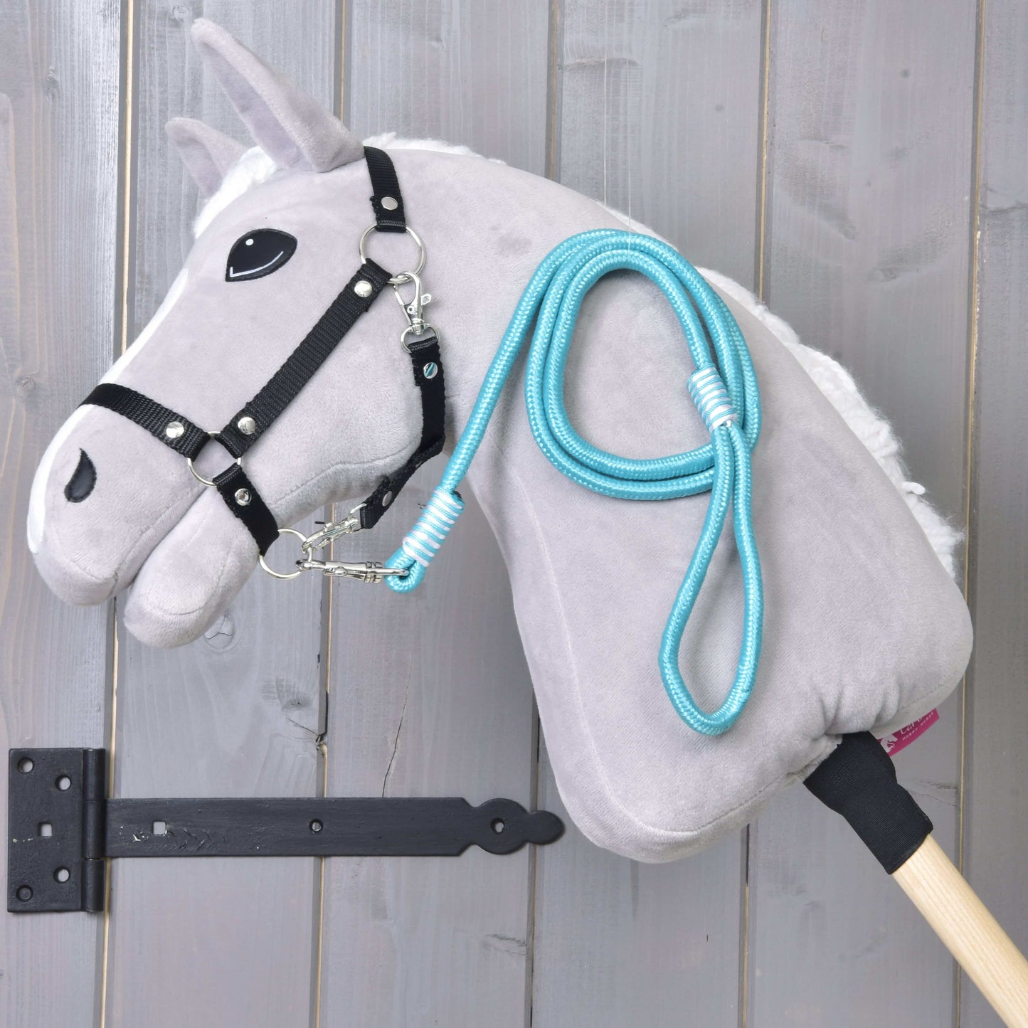 Hobby Horse - vodítko Atlantis od Hobby Horse LarDen s šedou plyšovou hlavou, černou uzdečkou a modrým provazovým vodítkem, upevněné na dřevěné tyči a zobrazené před svislými dřevěnými prkny.