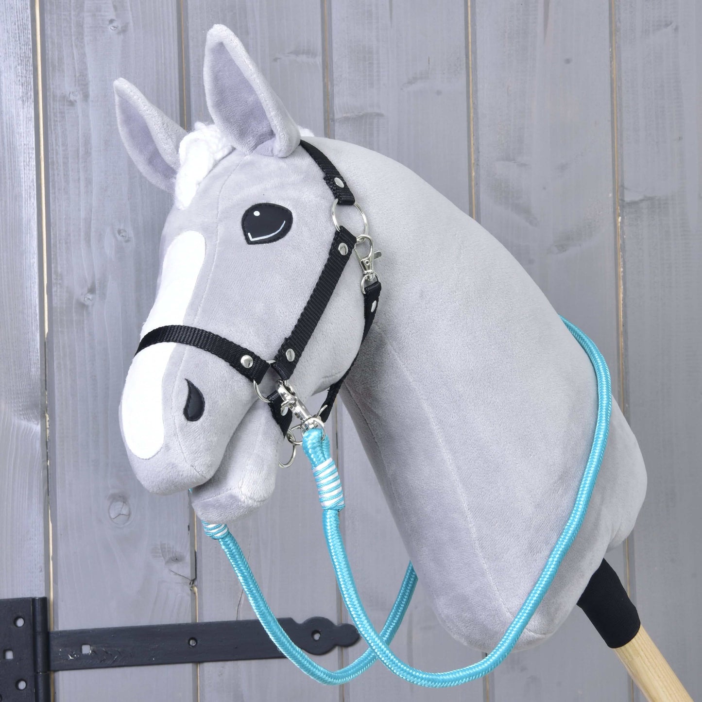 Hobby Horse LarDen Otěže Atlantis na šedém plyšovém Hobby Horse s černou uzdečkou a modrými polypropylenovými lanovými otěžemi, na dřevěné tyči před rustikální dřevěnou stěnou.