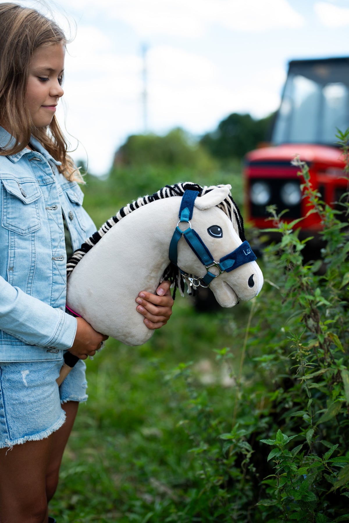 Dívka v džínové bundě drží u živého plotu Hobby Horse Aurora s černou ohlávkou od Hobby Horse LarDen, v pozadí je rozmazaný červený traktor, krémově bílá plyšová hlava, černobílá pruhovaná přízová hříva, s ohlávkou LarDen Blue s kožešinovým lemováním