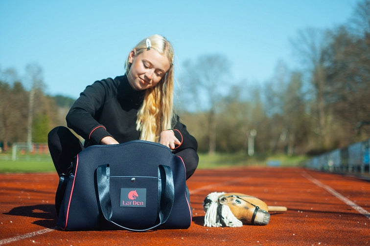 Mladá žena s dlouhými blond vlasy sedí na atletické dráze a dívá se do tmavě modré sportovní tašky s nápisem LarDen. Vedle ní na zemi leží hobby horse, naznačující její vášeň pro hobby horsing. V pozadí jsou vidět stromy a čáry atletické dráhy.