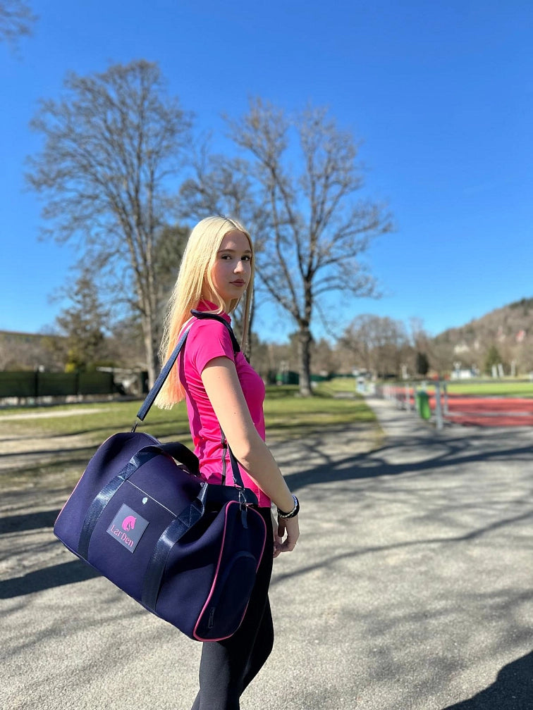 Mladá žena s dlouhými blond vlasy v růžovém sportovním tričku a s tmavě modrou sportovní taškou stojí venku za slunečného dne se svým hobby horse, připravená na zábavný den Hobby Horsing pod jasně modrou oblohou a mezi zelenými stromy.