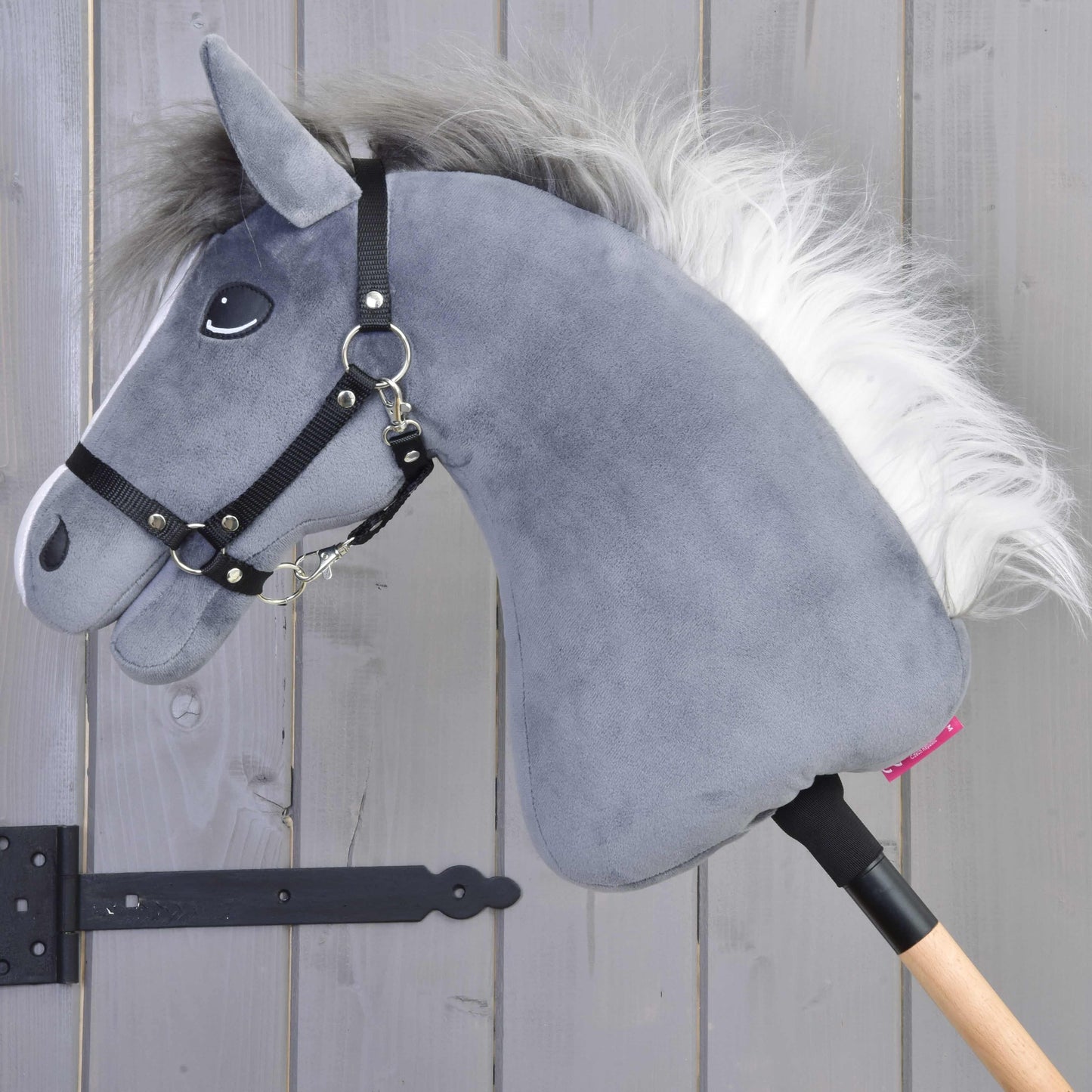Hobby Horse Beauty s černou ohlávkou od Hobby Horse LarDen je plyšová šedá hračka anglického plnokrevníka na dřevěné tyči s bílou hřívou a černou ohlávkou, na pozadí světle šedých dřevěných prken.