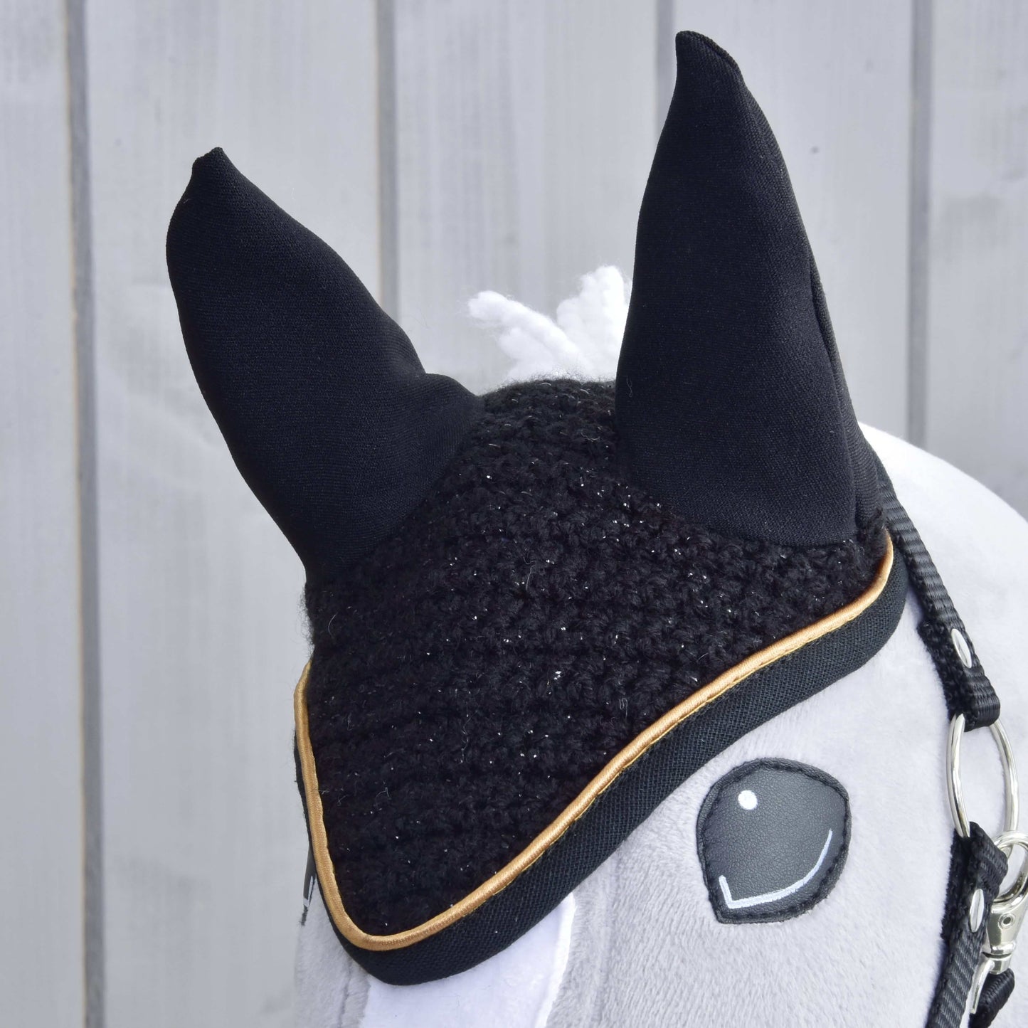 Detail plyšového koně od Hobby Horse LarDen s čabrakou háčkovanou Black Gold pro ochranu proti hmyzu. Kůň má uzdečku, bílou hřívu a je na pozadí světlé dřevěné stěny.