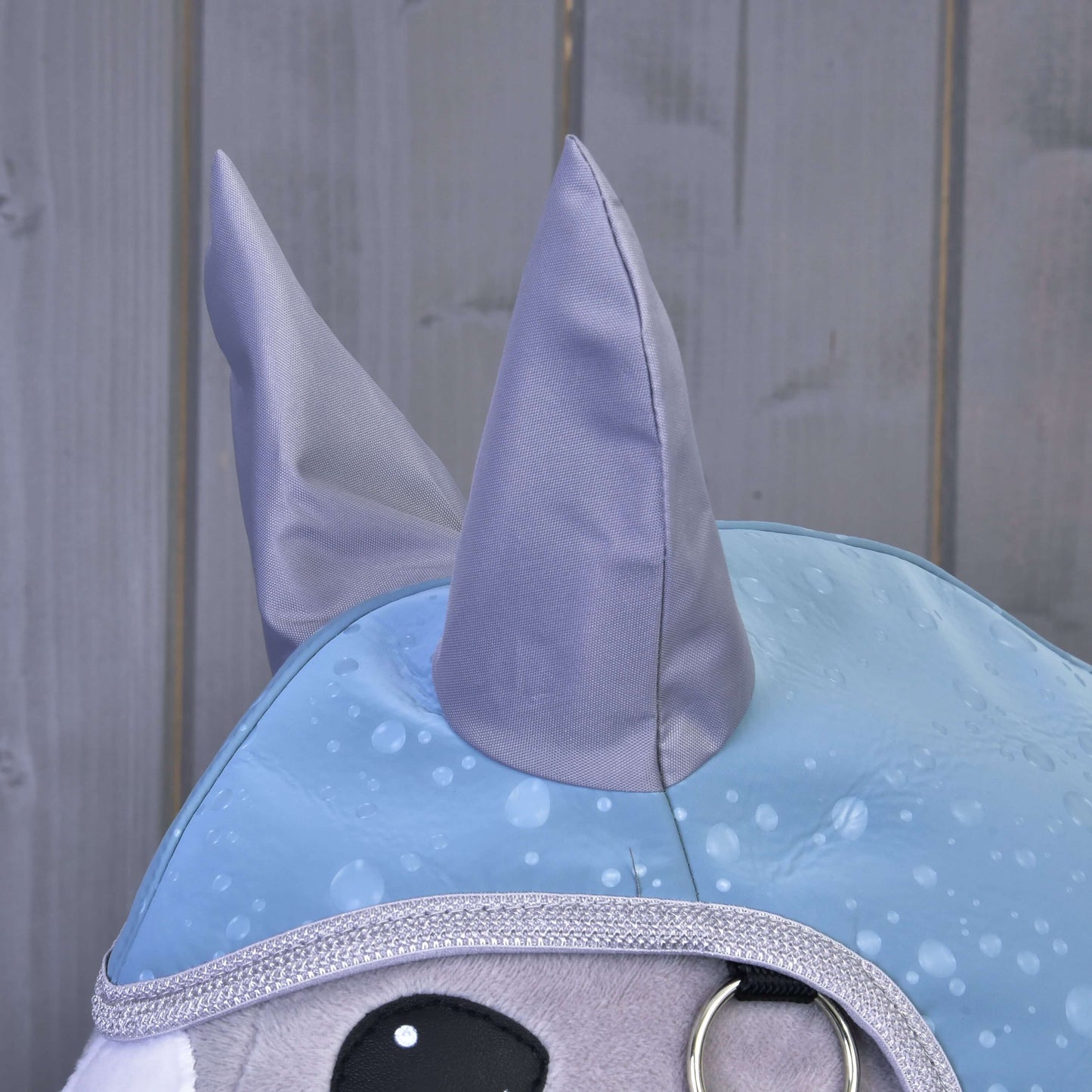 Detail plyšové hračky Hobby Horse LarDen v outfitu „Raincoat Blue Drops“ s lesklýma šedýma ušima, stříbrným lemováním, velkýma černýma očima a modrým pláštěm se vzorem kapek deště. V pozadí jsou hladké šedé dřevěné panely.