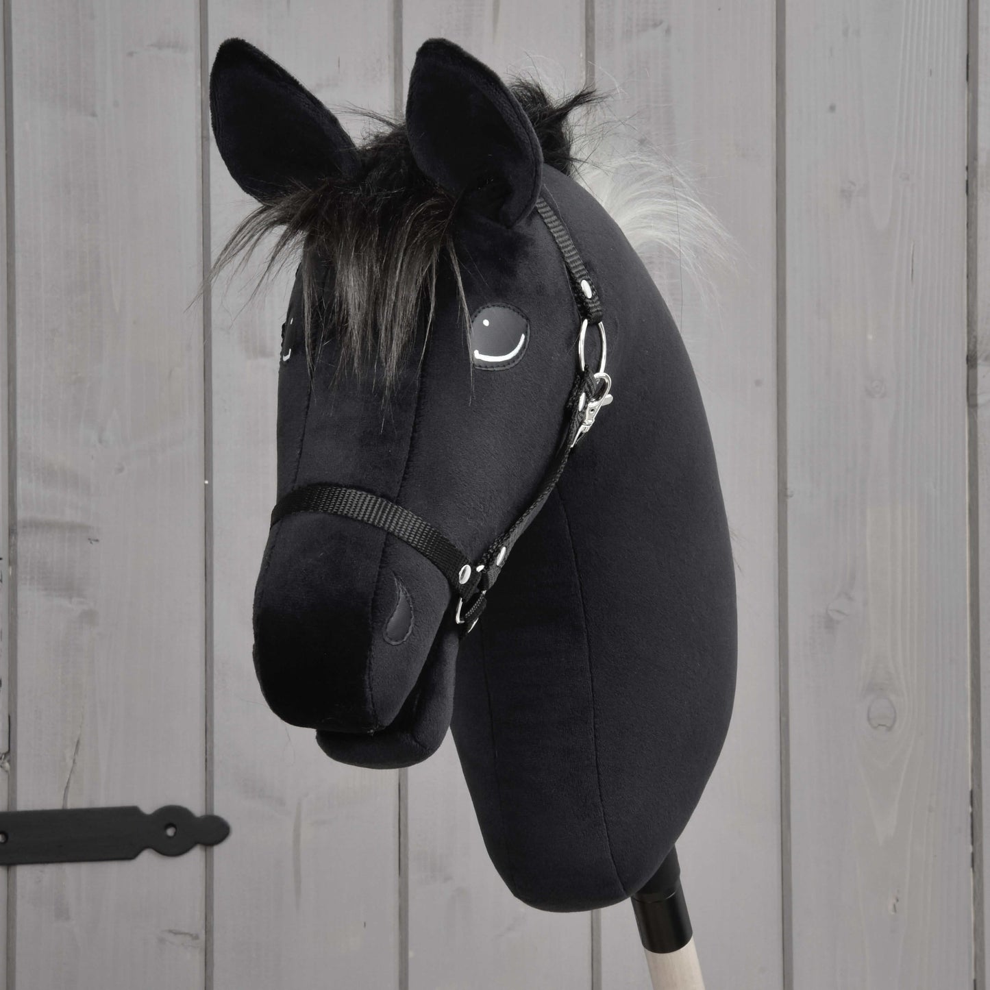 Hobby Horse Bond s černou ohlávkou od Hobby Horse LarDen, s černou plyšovou hlavou inspirovanou hannoverským koněm, bílou hřívou, vyšívanými detaily a černou ohlávkou, nasazený na dřevěné tyči před šedou dřevěnou stěnou.
