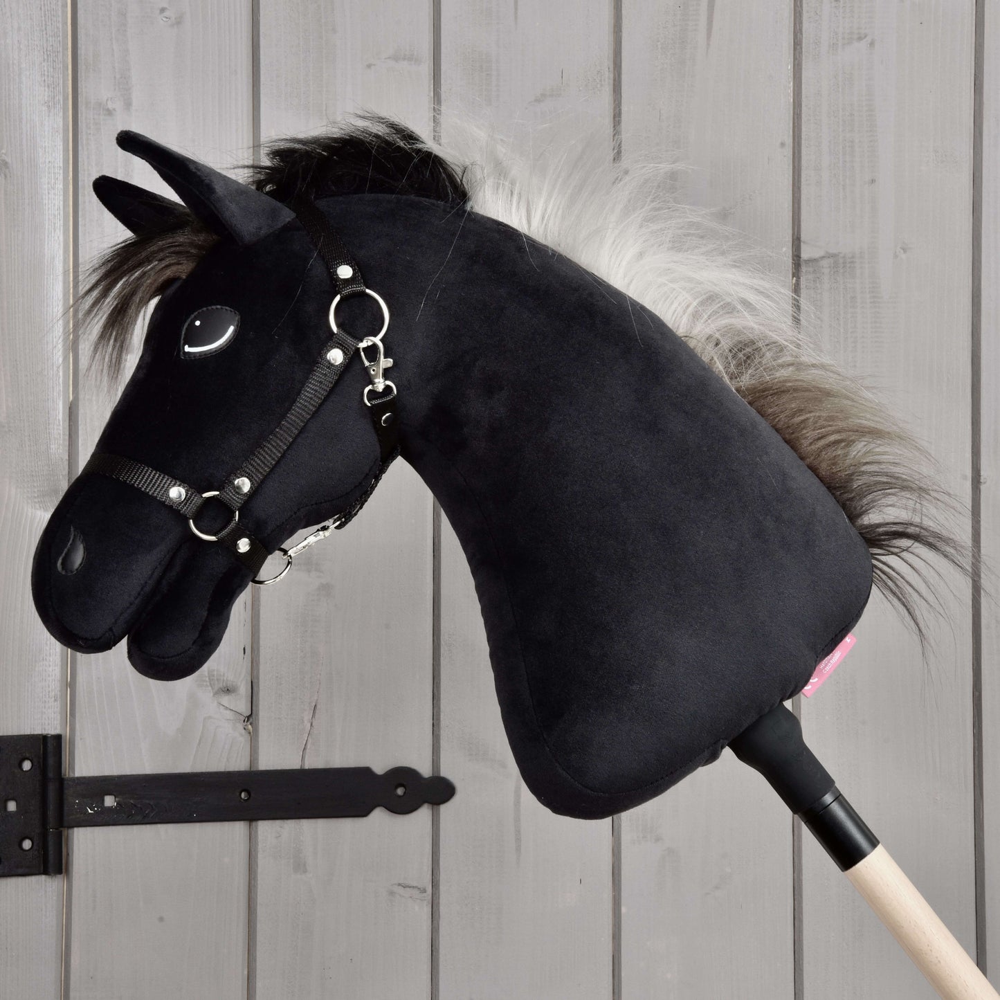 Hobby Horse Bond s černou ohlávkou od Hobby Horse LarDen, černý plyšový Hobby Horse s bílou hřívou, černou ohlávkou a otěžemi na dřevěné tyči, zobrazený před světlou dřevěnou stěnou – ideální pro každého fanouška Hobby Horse Bond.