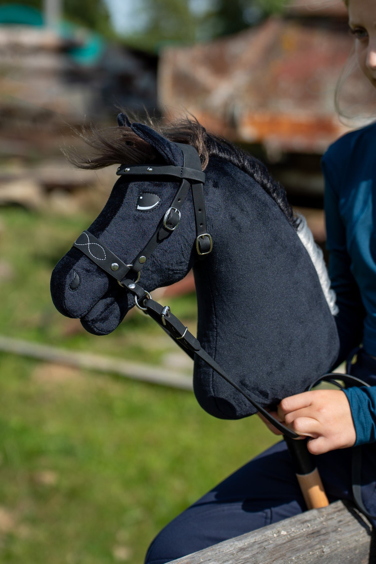 Hlava Hobby Horse Bond s černou ohlávkou od Hobby Horse LarDen před rozmazaným hospodářským dvorem, černý plyš, tmavě hnědá přízová hříva, černé řemínky se stříbrnými přezkami, se zdobenou bezudidlovou uzdečkou