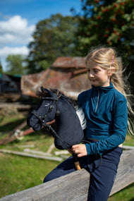 Blonďatá dívka v tyrkysovém topu sedí na dřevěné ohradě ve slunném hospodářském dvoře a drží Hobby Horse Bond s černou ohlávkou od Hobby Horse LarDen, černá plyšová hlava, tmavě hnědá přízová hříva, se zdobenou bezudidlovou uzdečkou