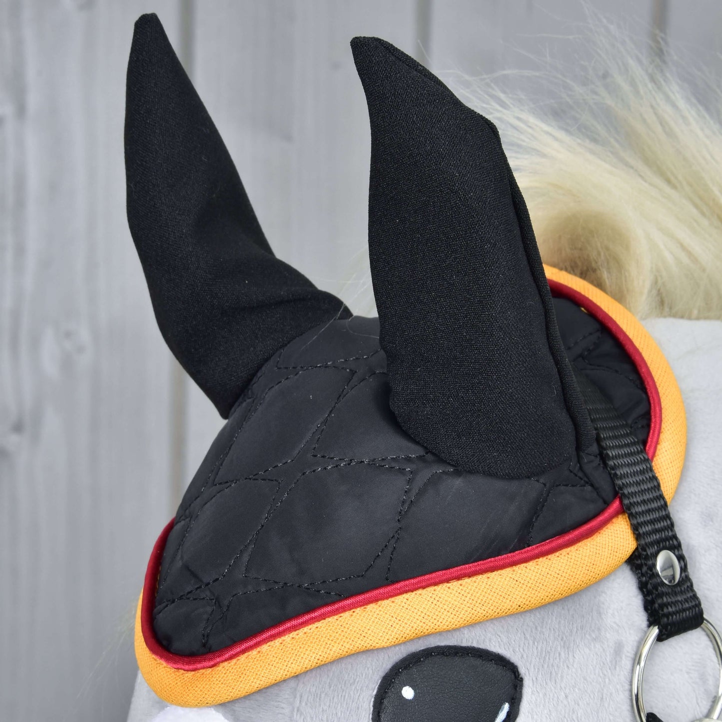 Detail koně s čabrakou Hobby Horse LarDen Flag DE, černou prošívanou čabrakou se žlutým a červeným lemováním k ochraně proti hmyzu, zakrývající uši a část hlavy na světlém dřevěném pozadí.