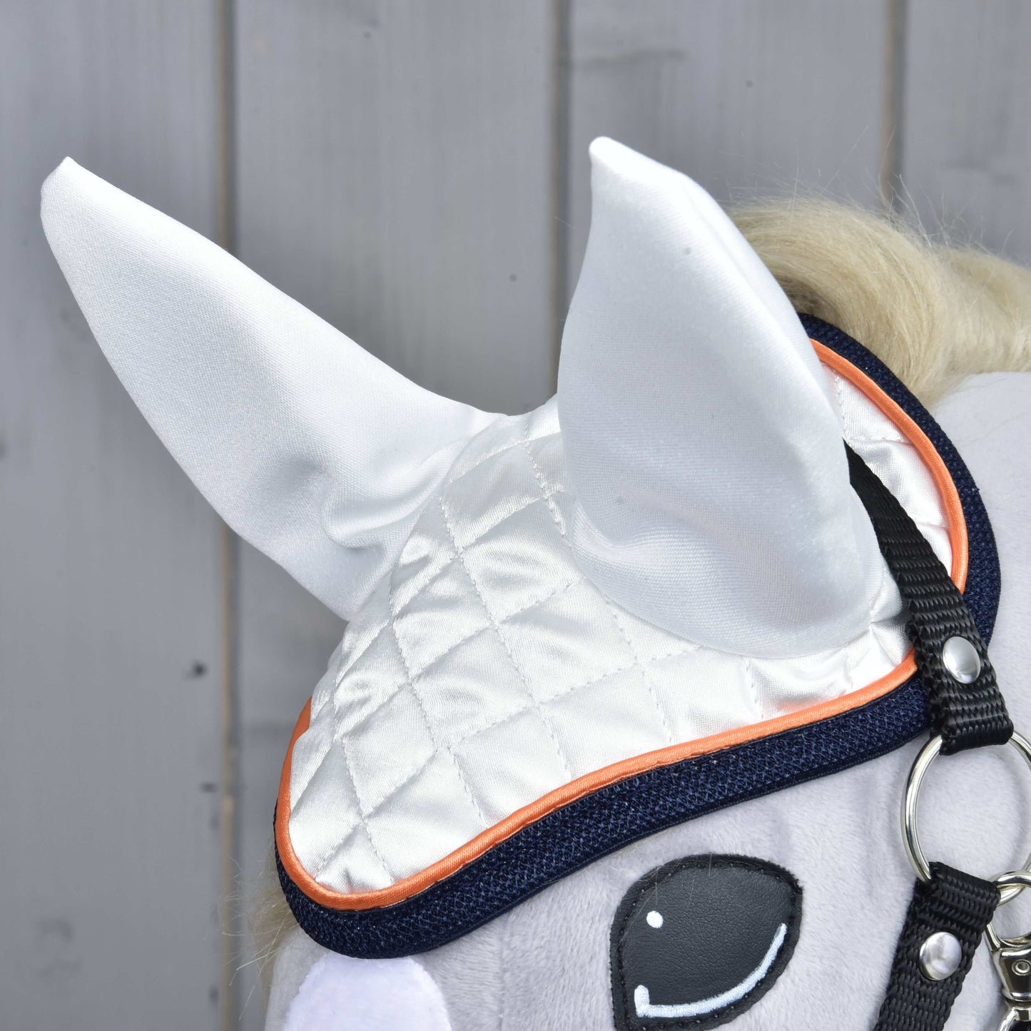Čabraka Speedy od Hobby Horse LarDen, detail v bílé barvě s oranžovým lemováním, vystavená na plyšové hračce hobby horse s viditelným levým okem a uzdečkou – stylová ochrana proti mouchám na dřevěném pozadí.
