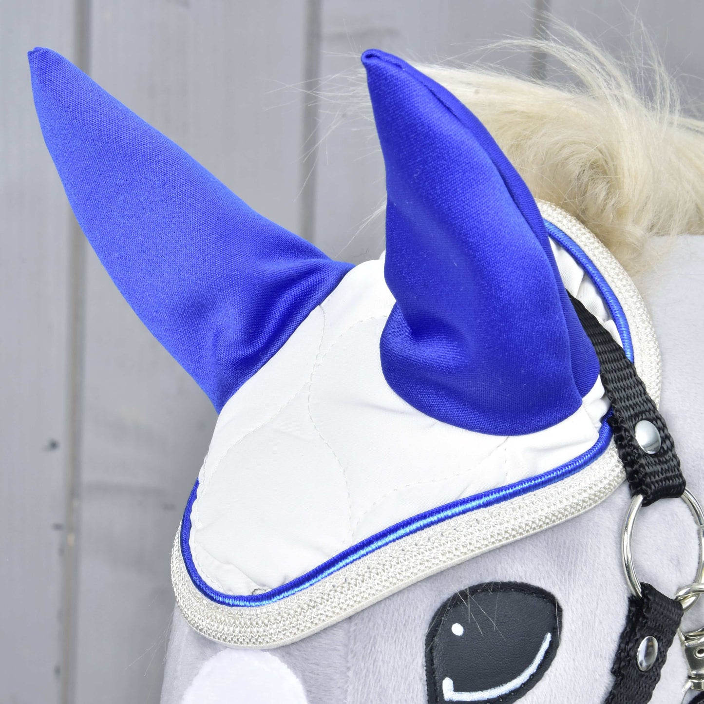 Detail hlavy plyšové hračky Hobby Horse LarDen s bílým látkovým obličejem, modrýma ušima, černou ohlávkou a páskou přes oko. Hračka má na sobě čabraku Cream Pearl k ochraně proti hmyzu na pozadí šedého dřeva.