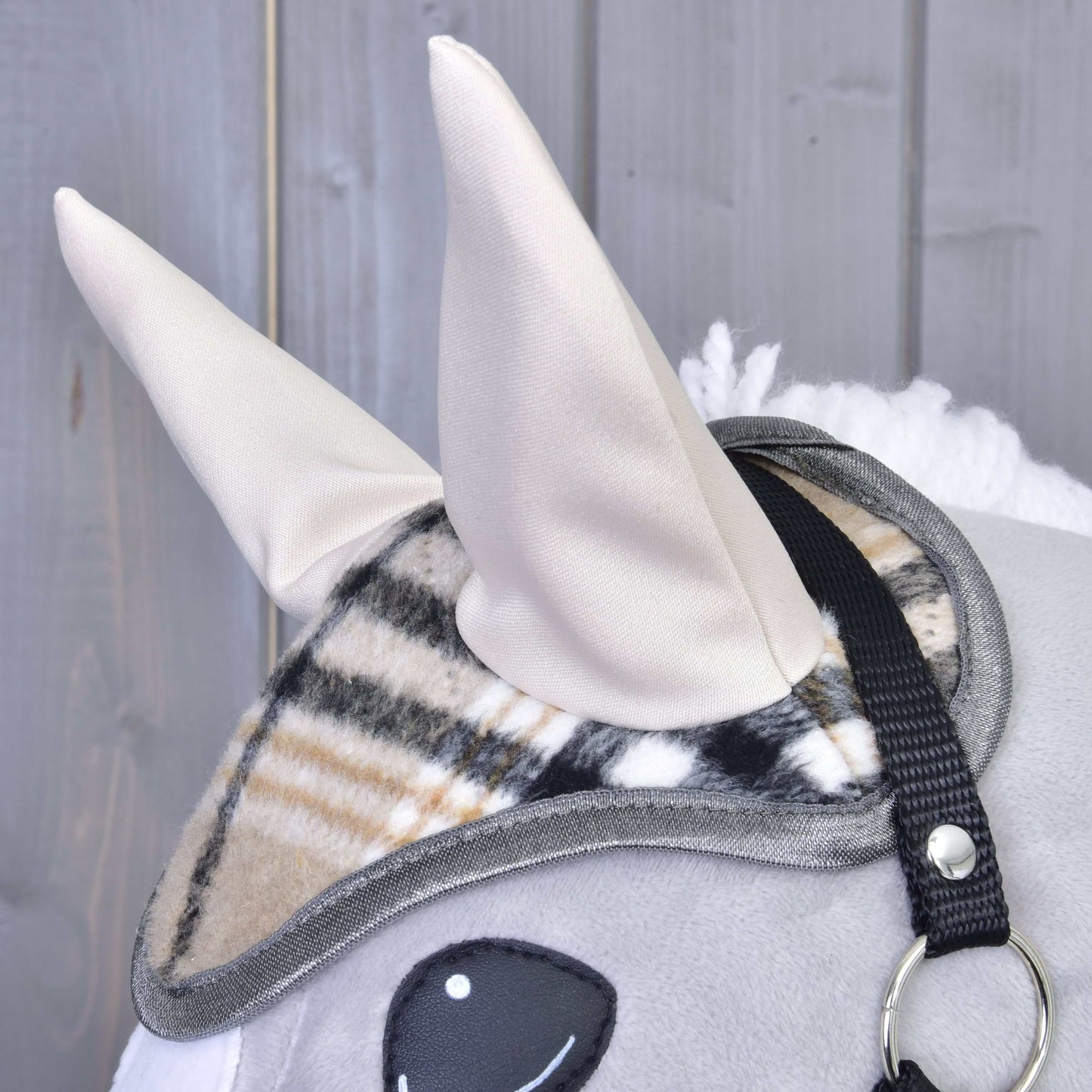 Detail plyšového Hobby Horse od Hobby Horse LarDen se dvěma béžovými látkovými ušima, s čabrakou Cremo England s kostkovaným vzorem pro ochranu před mouchami a černým řemínkem uzdečky, na šedém dřevěném pozadí.