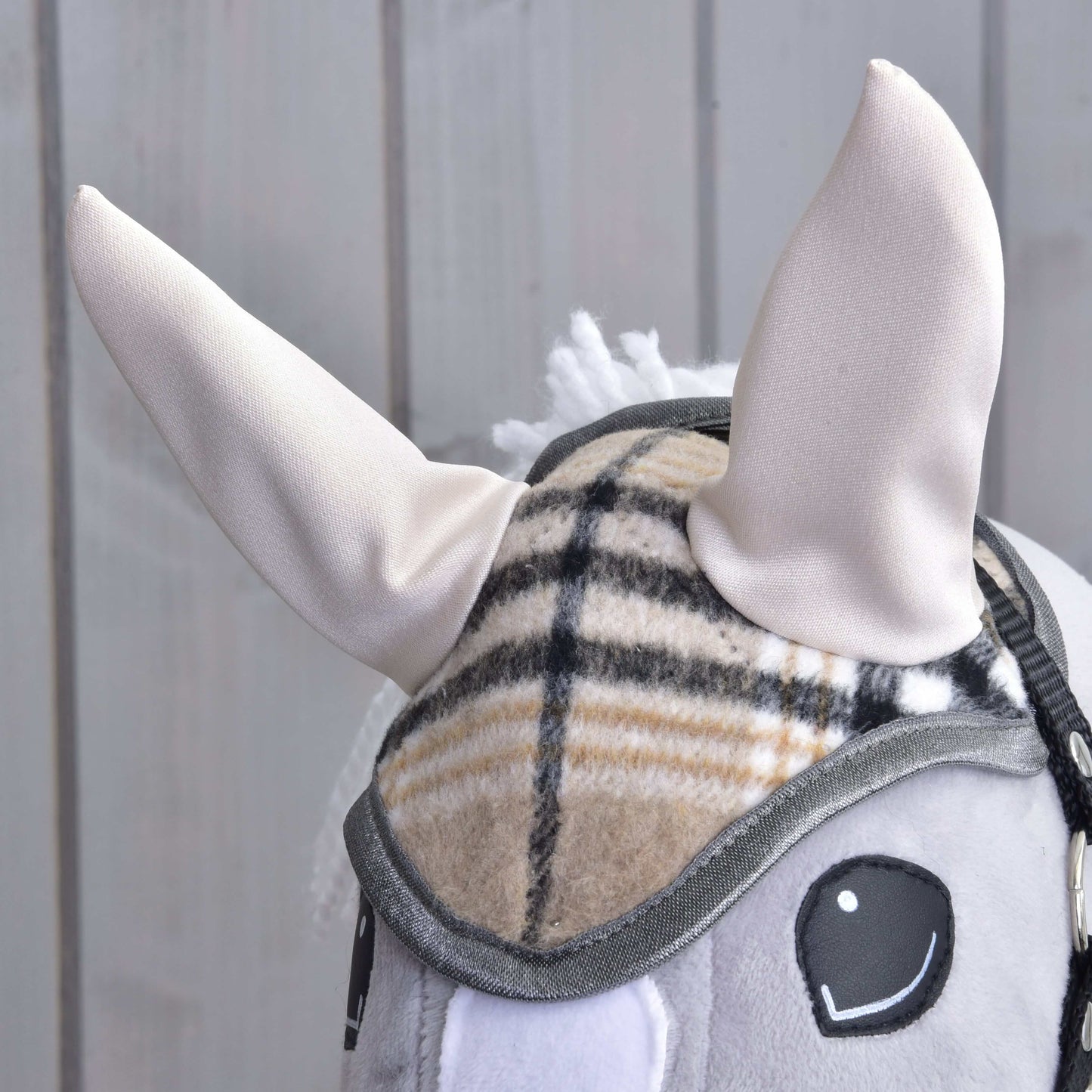 Detail Hobby Horse LarDen Čabraka Cremo England: má měkké krémově zbarvené uši, kostkovaný řemínek uzdečky pro ochranu před mouchami a šedou hřívu, zobrazená na světlém dřevěném pozadí.