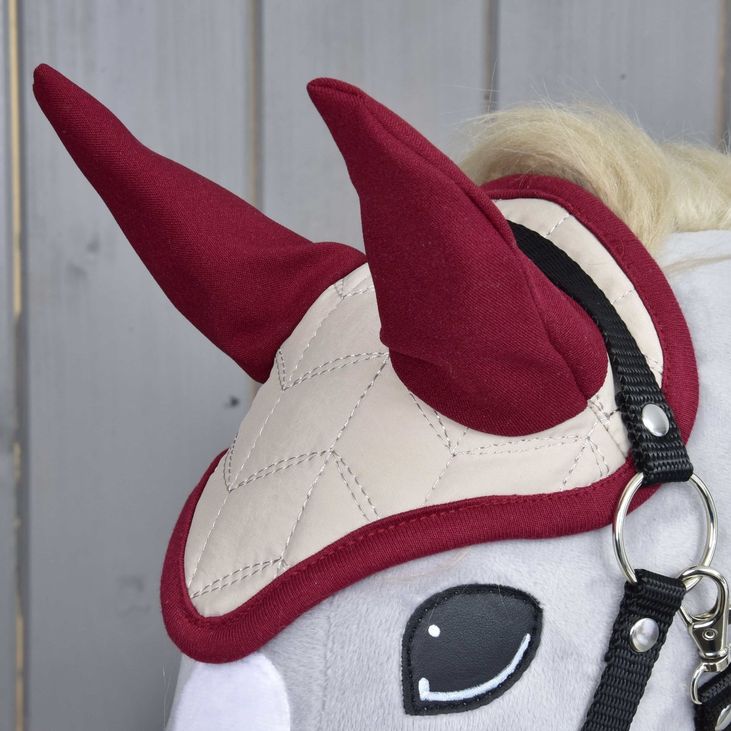 Detail Hobby Horse LarDen s čabrakou Maroon Star, s béžovým prošíváním pro ochranu proti hmyzu, dvěma vzpřímenými červenými tvary připomínajícími rohy a černou uzdečkou. V pozadí jsou světlé dřevěné panely.