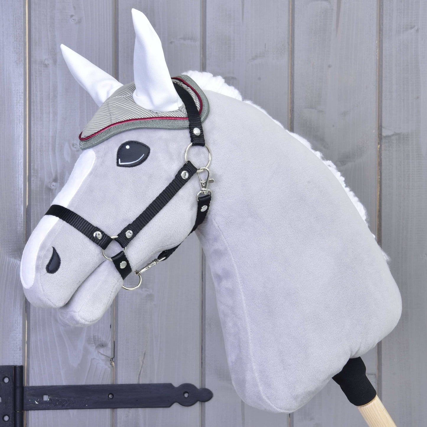 Detail šedého plyšového Hobby Horse od Hobby Horse LarDen s uzdečkou, ozdobným čelenkovým řemínkem a čabrakou Silver Eleganza pro ochranu před hmyzem, na pozadí dřevěného plotu.