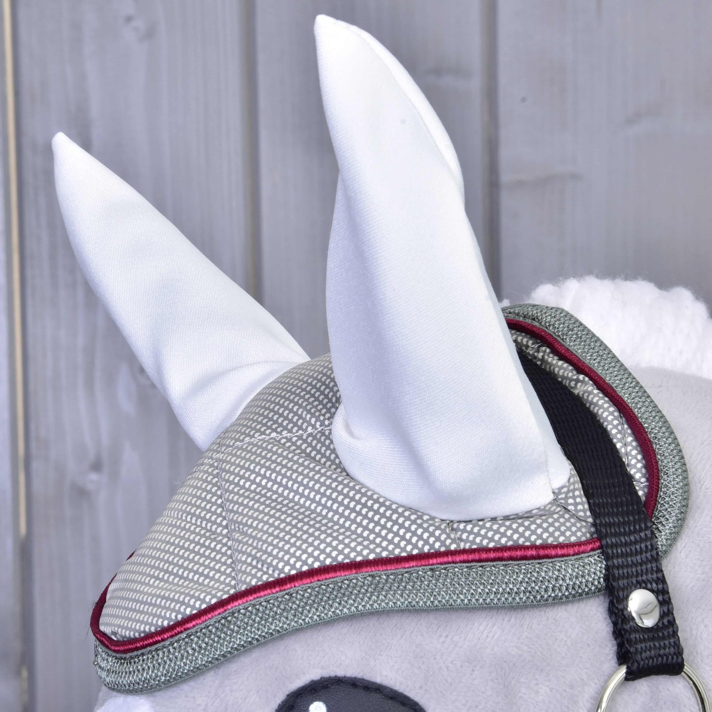 Detail šedého Hobby Horse od Hobby Horse LarDen s čabrakou Silver Eleganza a šedo-červenou uzdečkou, s velkými bílými ušima, na pozadí dřevěných panelů.