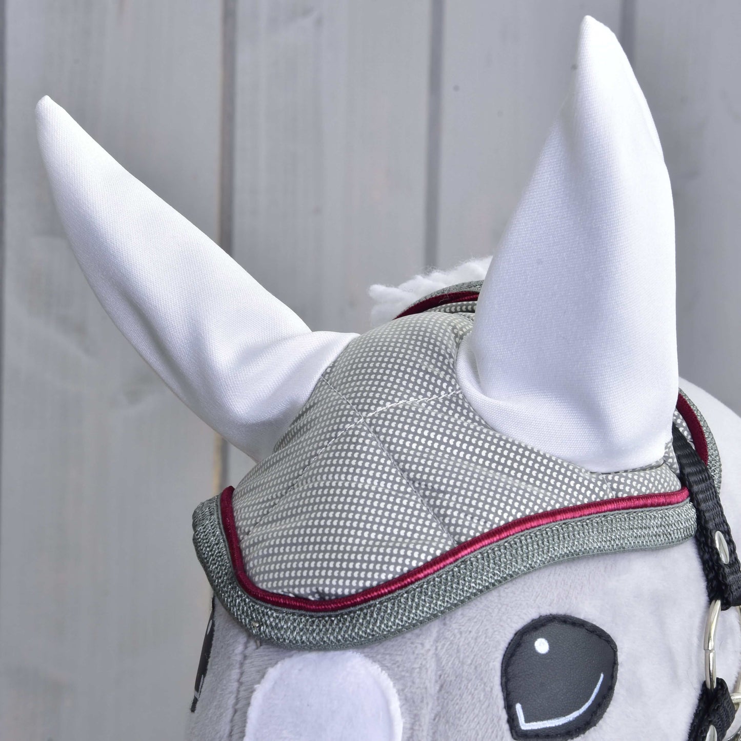 Detail Hobby Horse LarDen Čabraka Silver Eleganza: šedá síťovaná čabraka s červeným lemováním, velkými vztyčenými bílými ušima a černě vyšívanýma očima, ideální pro ochranu před hmyzem, na světlém dřevěném pozadí.