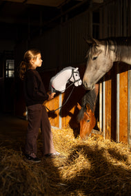 Tmavovlasá dívka v černé mikině stojí v uličce stáje a drží Hobby Horse Caesar s černou Ohlávkou od Hobby Horse LarDen se šedou plyšovou hlavou a černobílou hřívou z příze, zatímco z boxů vyhlížejí šedý kůň a hnědák, s kompletní zdobenou bezudidlovou uzdečkou
