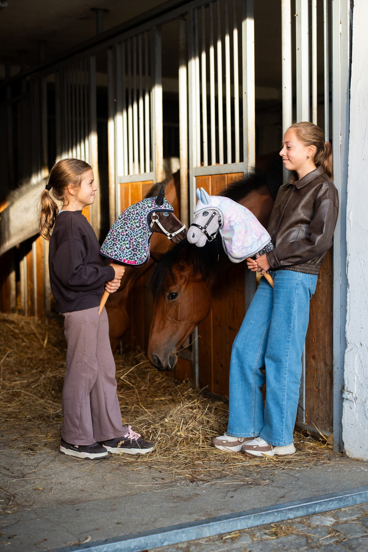 Lifestyle photo: two girls at the stable door, one holding the Hobby Horse Caesar with Black Halter in the Raincoat Palete and the other the Hobby Horse Alfredo with Black Halter in the Raincoat Confetti (softshell), a bay horse looking out of its box behind them - both by Hobby Horse LarDen, shown here with the Complete bitless bridle, decorated.