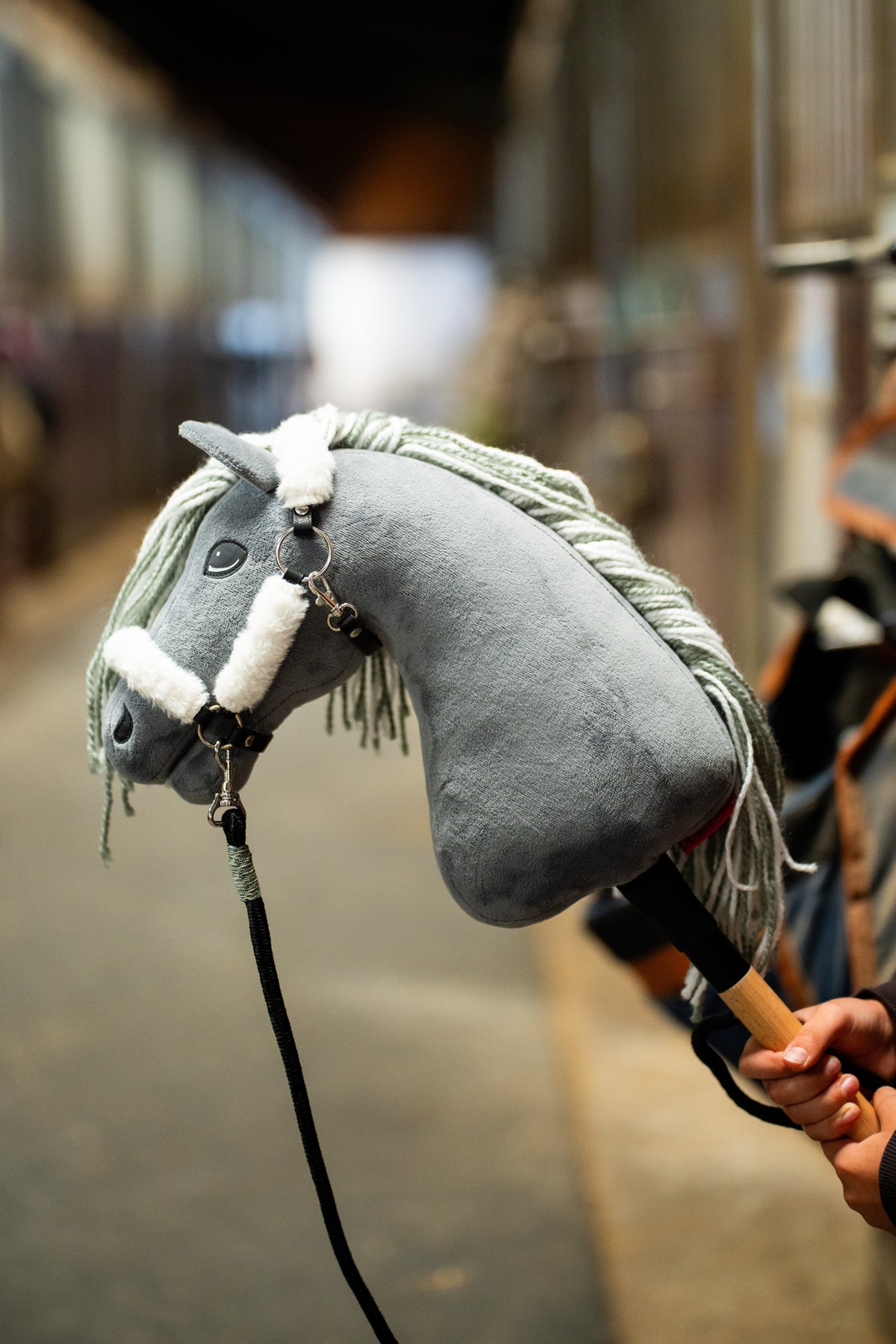 Hlava Hobby Horse Calypso s černou ohlávkou od Hobby Horse LarDen držená v uličce stáje před rozmazanými boxy, šedý plyš, světle šedá přízová hříva, tmavé oko, s ohlávkou Teddy Classic s kožešinovým lemováním a černým vodítkem