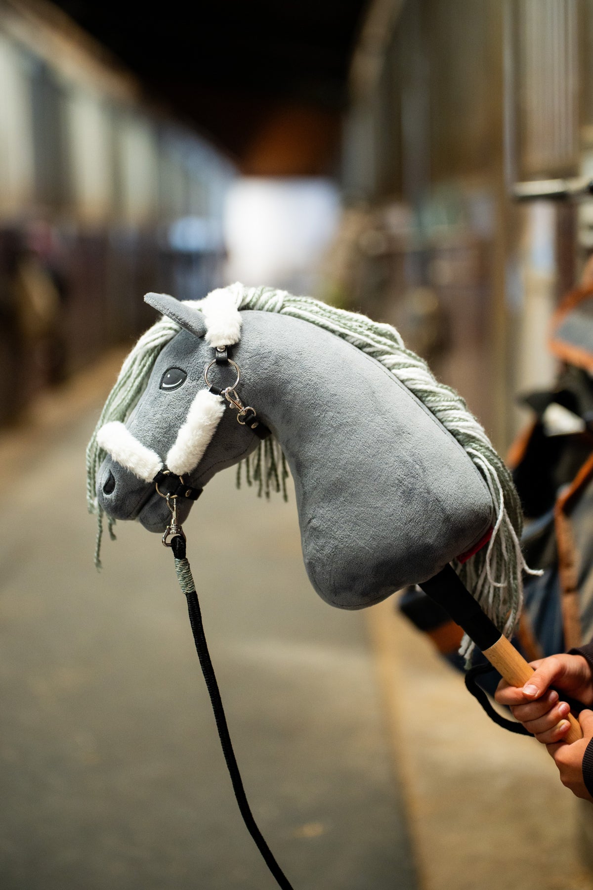 Hlava Hobby Horse Calypso s černou ohlávkou od LarDen je zvednutá v uličce stáje, za ní jsou rozmazané boxy, s šedým plyšem, světle šedou přízovou hřívou a tmavým okem, s ohlávkou Teddy Classic podšitou kožíškem a černým vodítkem