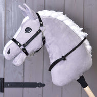 Nákrčník černý od Hobby Horse LarDen na šedém plyšovém Hobby Horse s bílou hřívou na dřevěné tyči, s černou uzdečkou, otěžemi a ručně vyrobeným cordeem, před šedou dřevěnou stěnou.