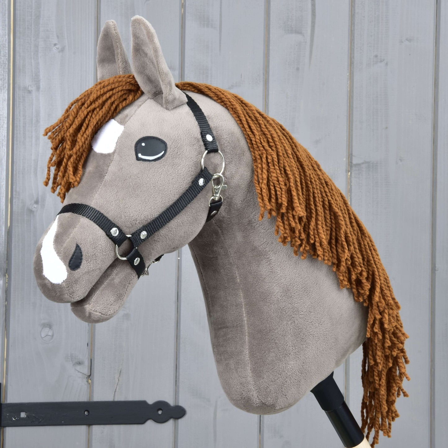 Hobby Horse Chanell s černou ohlávkou od Hobby Horse LarDen, s měkkým plyšovým provedením, černou ohlávkou, bílými znaky na hlavě, hnědou přízovou hřívou a kreslenýma očima, zobrazený před světle šedou dřevěnou stěnou.