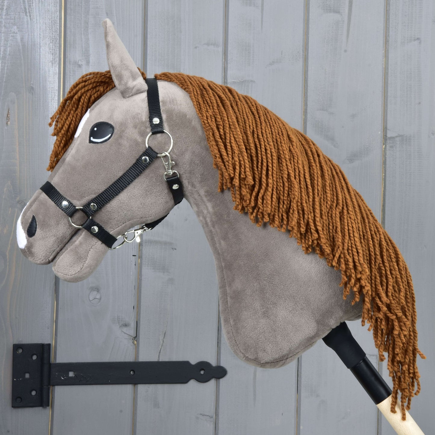 Hobby Horse Chanell s černou ohlávkou od Hobby Horse LarDen, se šedou látkovou hlavou, hnědou přízovou hřívou, černou ohlávkou a bílými plstěnými detaily, nasazený na dřevěné tyči před stěnou z šedých dřevěných prken.
