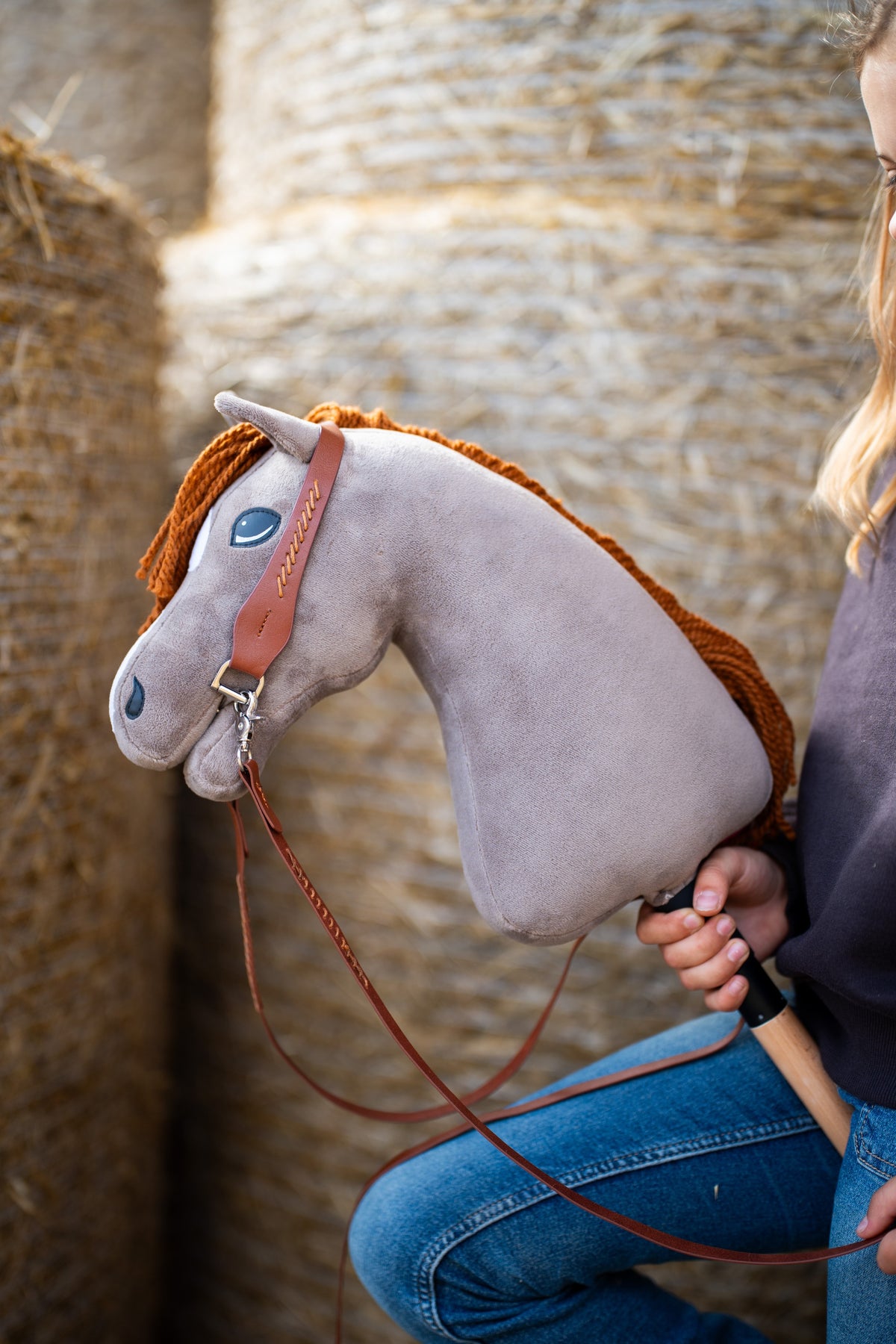 Hlava Hobby Horse Chanell s černou ohlávkou od Hobby Horse LarDen před kulatými balíky slámy, šedý plyš, modré oko, zrzavá přízová hříva, s kompletní westernovou uzdečkou s jedním náušníkem z hnědé kůže