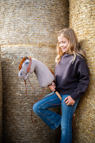 Usmívající se dívka v tmavě hnědé mikině se opírá o kulaté balíky slámy a drží Hobby Horse Chanell s černou ohlávkou od Hobby Horse LarDen, šedá plyšová hlava, modré oko, zrzavá přízová hříva, s kompletní westernovou uzdečkou s jedním náušníkem