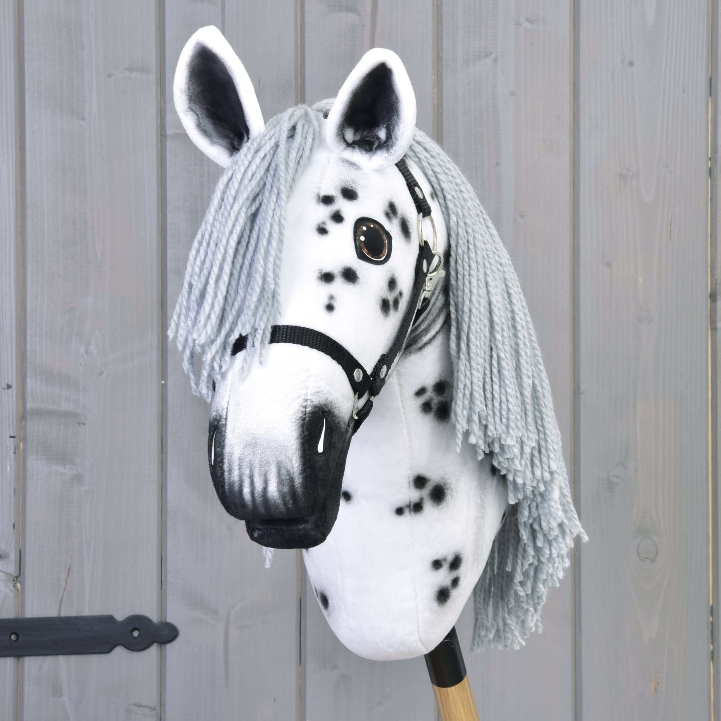 Hobby Horse Chantal s černou ohlávkou od Hobby Horse LarDen je ručně vyrobená klisna inspirovaná plemenem knabstrupper, s bílým obličejem, černými skvrnami, dlouhou šedou hřívou a na dřevěné tyči před šedou stěnou.