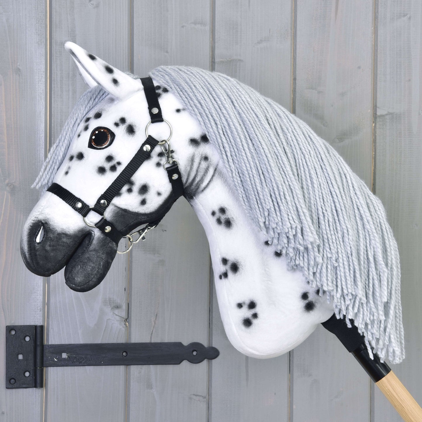 Hobby Horse Chantal s černou ohlávkou od Hobby Horse LarDen, s bílou srstí s černými skvrnami, šedou přízovou hřívou a černou ohlávkou. Tento ručně vyrobený Hobby Horse ve stylu knabstruppera je na dřevěné tyči a naaranžovaný před šedou dřevěnou stěnou.
