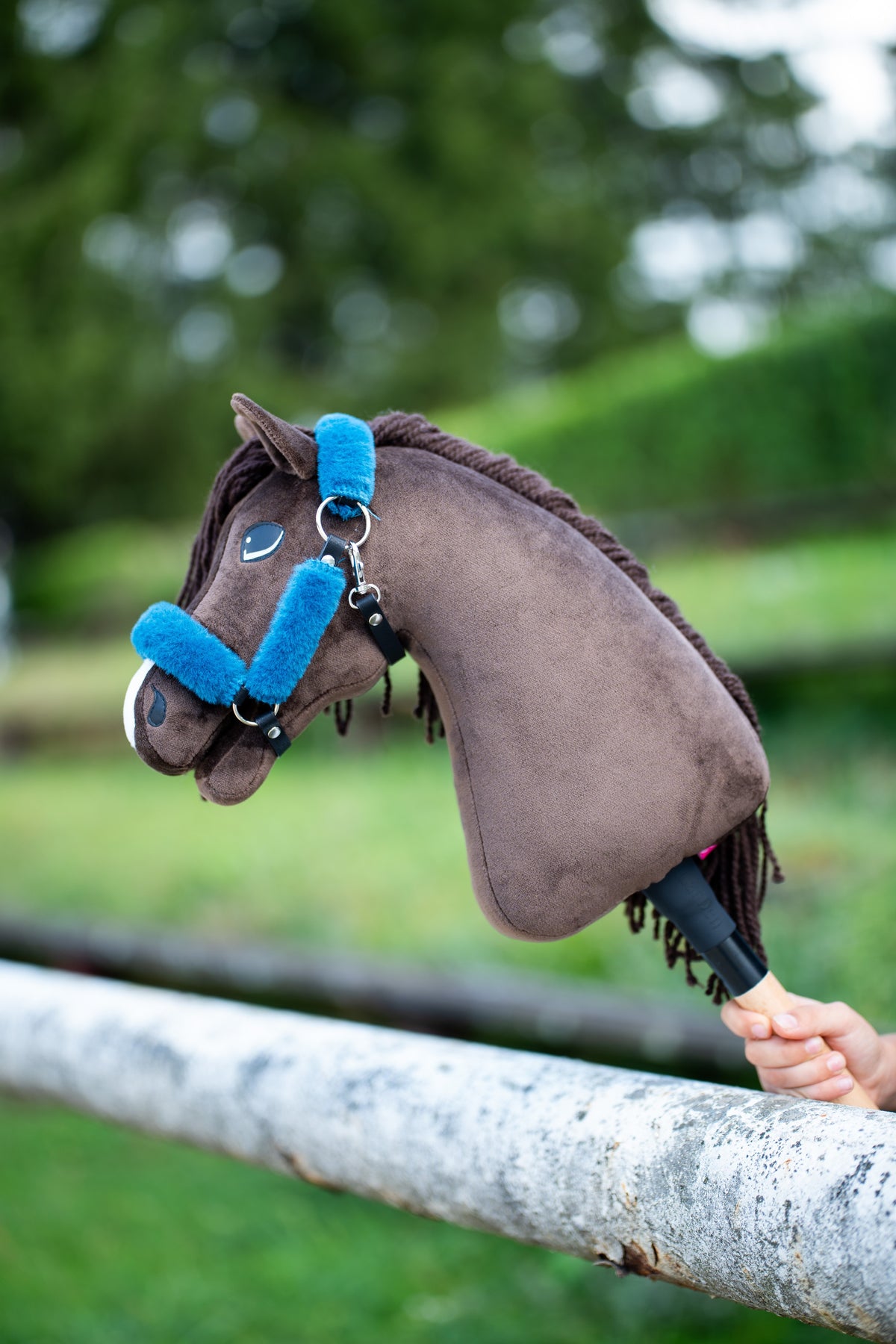 Hlava Hobby Horse Chianti s černou ohlávkou od Hobby Horse LarDen spočívá přes bílou ohradu, tmavě hnědý plyš, tmavá přízová hříva a ofina, s ohlávkou Teddy Petrol s kožešinovým lemováním v jasně modré barvě