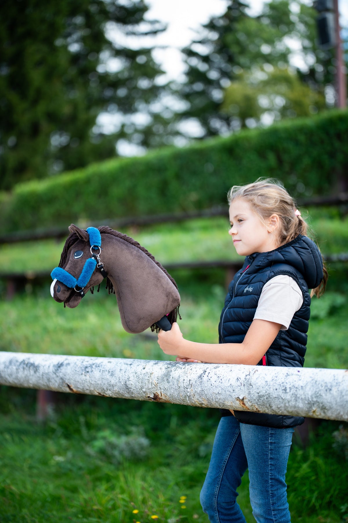 Dívka v tmavomodré prošívané vestě stojí za bílou ohradou a drží Hobby Horse Chianti s černou ohlávkou od Hobby Horse LarDen, za ní je zastřižený zelený živý plot, tmavě hnědá plyšová hlava, tmavá přízová hříva, s ohlávkou Teddy Petrol s kožešinovým lemováním