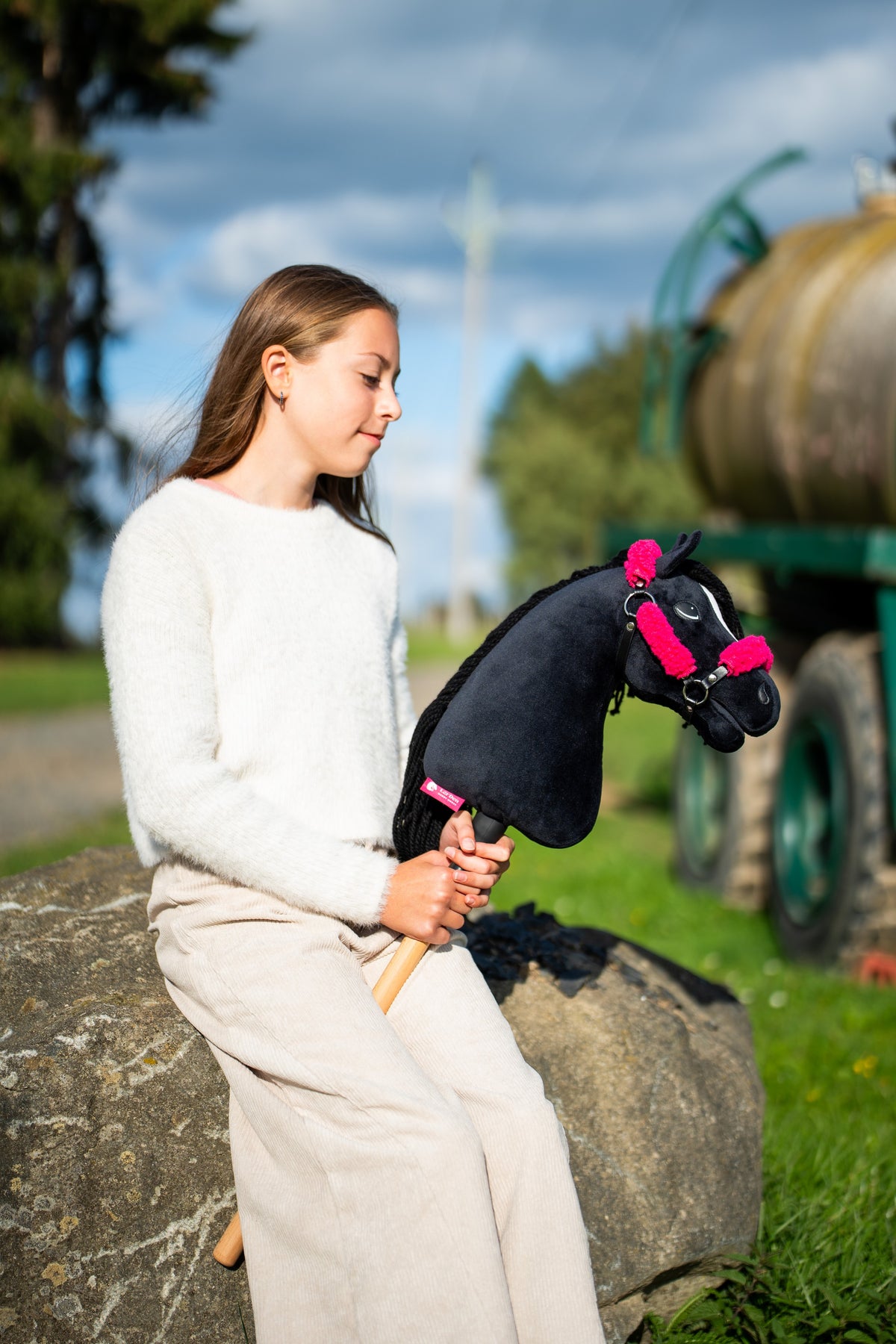 Dívka v krémovém svetru sedí na balvanu u cesty podél louky a drží Hobby Horse Cloony s černou ohlávkou od Hobby Horse LarDen, černá plyšová hlava, bílá hvězda, černá přízová hříva, s černou ohlávkou z koženky s kompletní růžovou kožešinou