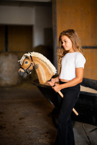 Dívka v bílém topu stojí ve stáji a drží Hobby Horse Colby s černou ohlávkou od Hobby Horse LarDen, plyšová hlava palomino, hustá krémová přízová hříva, černá ohlávka se stříbrným kováním