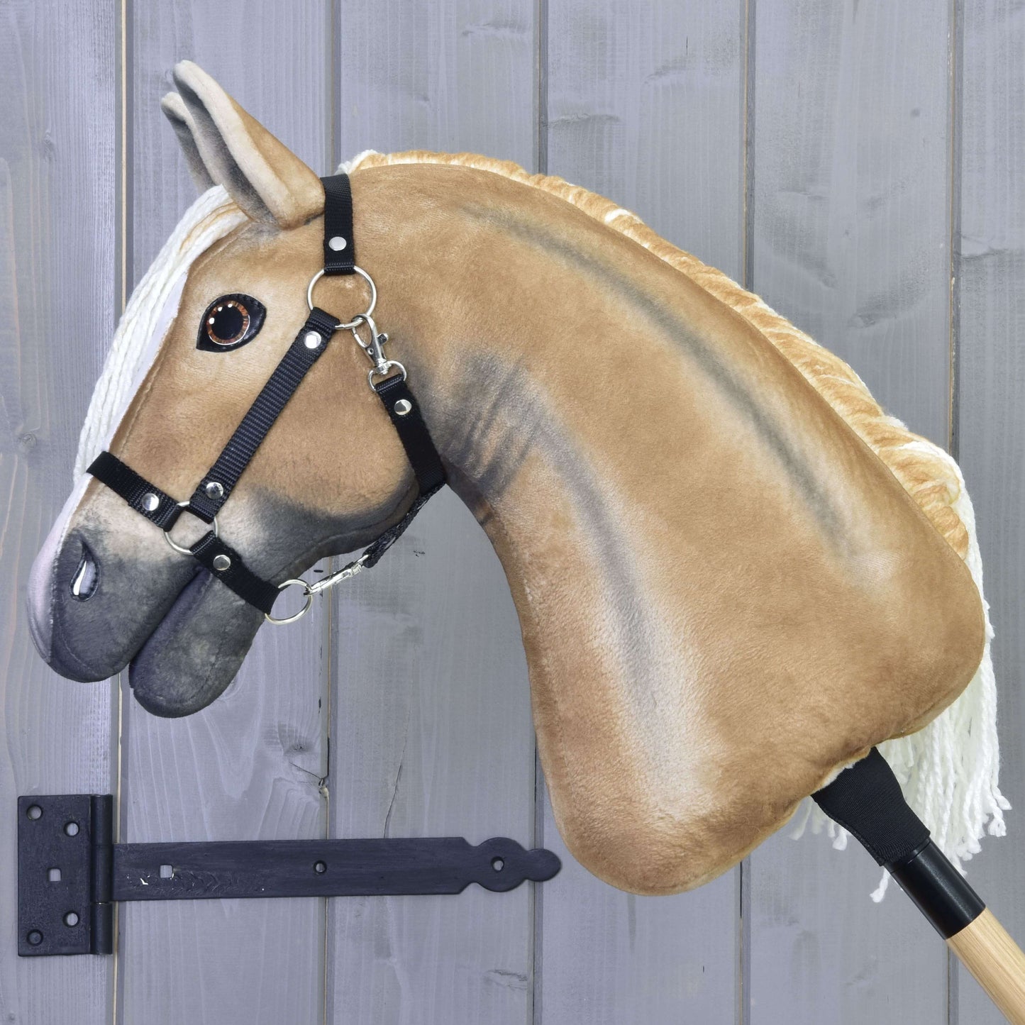 Hobby Horse Connie s černou ohlávkou od Hobby Horse LarDen s plyšovým béžovým tělem, bílou hřívou a černou ohlávkou, nasazený na dřevěné tyči před šedou dřevěnou stěnou.
