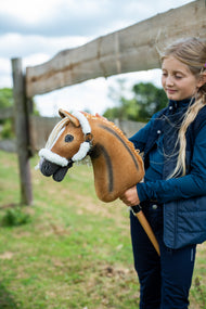 Dívka v tmavomodré vestě stojí u dřevěné ohrady a drží Hobby Horse Connie s černou ohlávkou od Hobby Horse LarDen, ryzá plyšová hlava s tmavým hřbetním pruhem, krémová přízová hříva, s ohlávkou Teddy Classic s kožešinovým lemováním