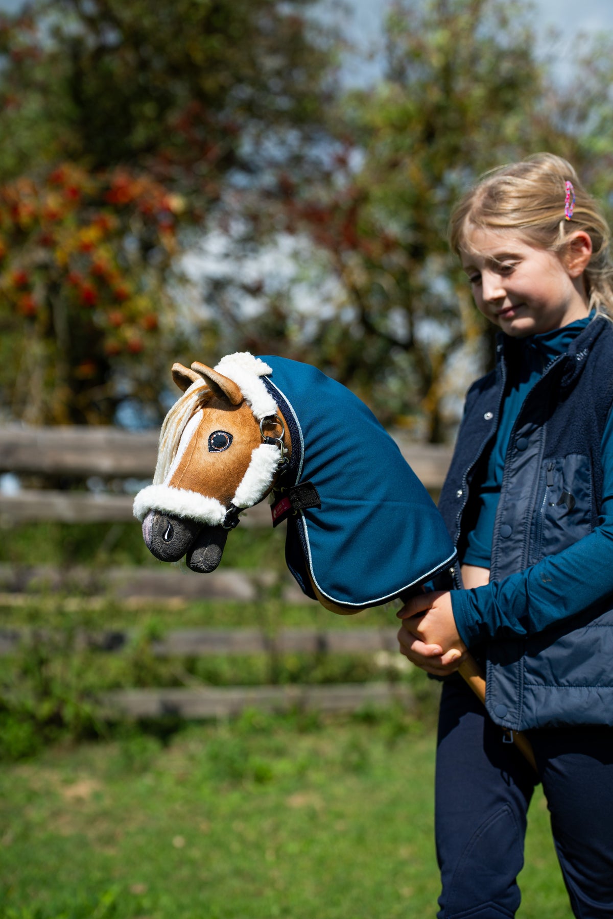 Dívka v tmavomodré vestě drží Hobby Horse Connie s černou ohlávkou od Hobby Horse LarDen v petrolejově modré stájové dece, v pozadí jsou podzimní bobule, ryzá plyšová hlava, krémová přízová hříva, s ohlávkou Teddy Classic s kožešinovým lemováním a stájovou dekou Winter Sky