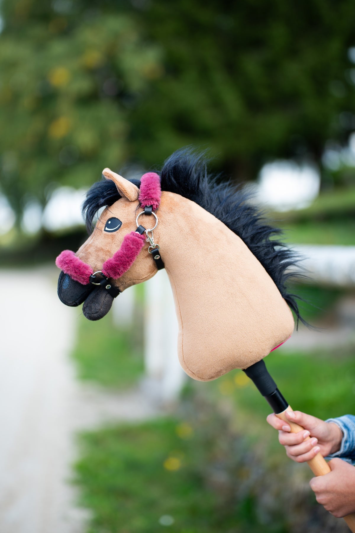 Hlava Hobby Horse Cookie s černou ohlávkou od Hobby Horse LarDen držená u cesty lemované stromy, pískový plyš, dlouhá černá přízová hříva, s ohlávkou Teddy Maroon s kožešinovým lemováním