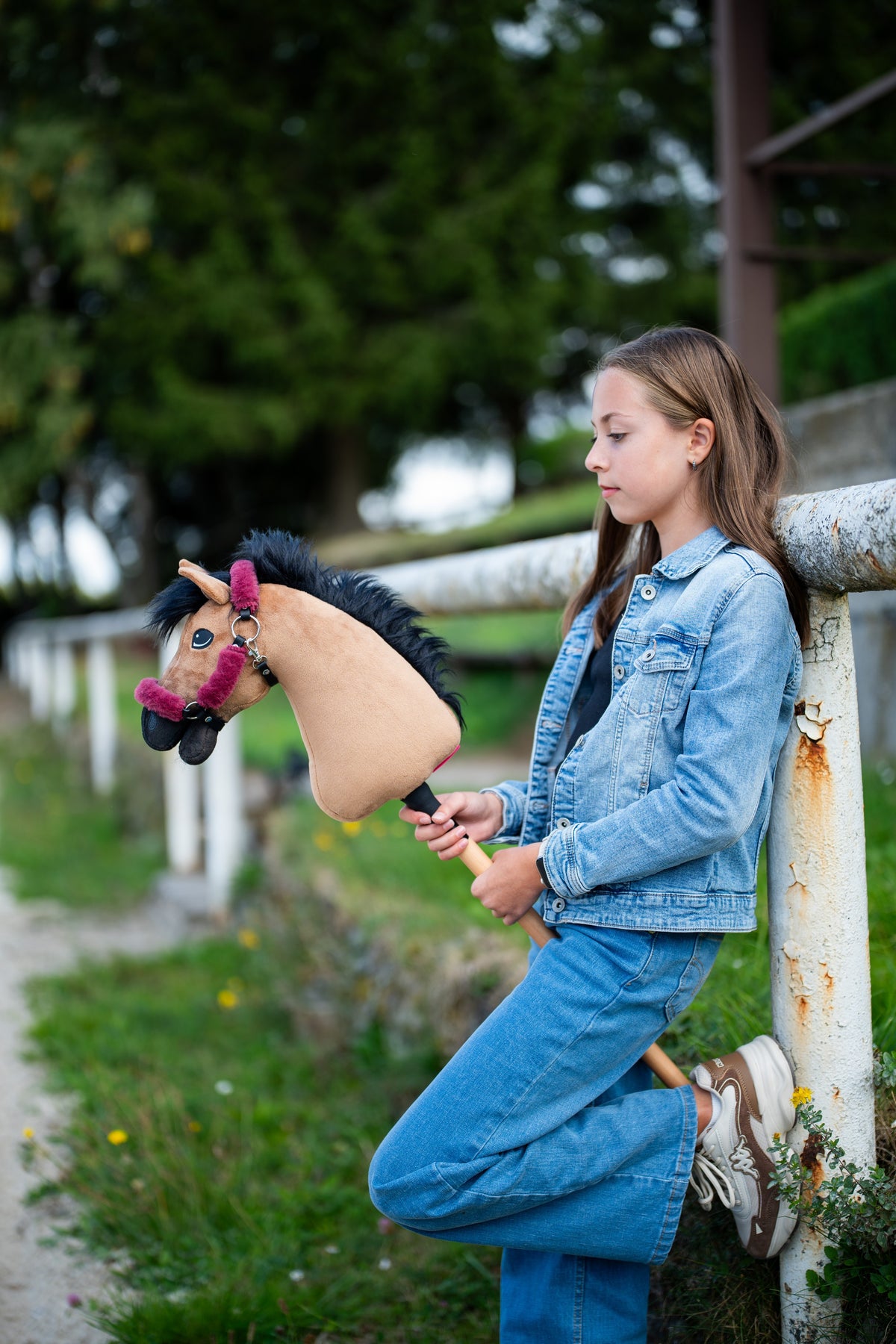 Dívka v džínovině se opírá o bílou ohradu a drží Hobby Horse Cookie s černou ohlávkou od Hobby Horse LarDen, písková plyšová hlava, černá přízová hříva, s ohlávkou Teddy Maroon s kožešinovým lemováním