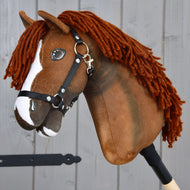 Hobby Horse Cooper s černou ohlávkou od Hobby Horse LarDen, hnědý Hobby Horse inspirovaný oldenburským sportovním koněm. Tato hračka má bílou lysinu na obličeji, hustou tmavě hnědou přízovou hřívu a černou nylonovou ohlávku s kovovými detaily. Je připevněná k dřevěné tyči a zobrazená před šedými dřevěnými prkny, ideální pro kreativní drezurní hru.