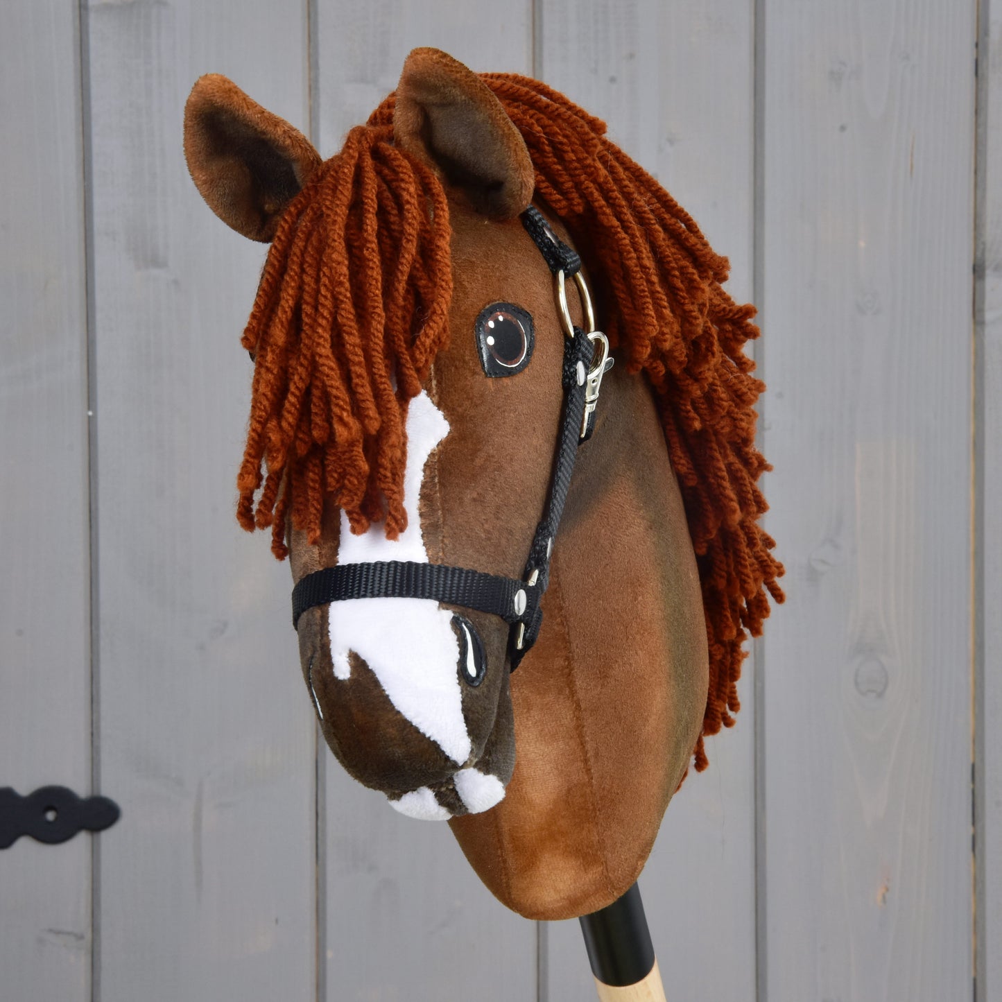 Hobby Horse Cooper s černou ohlávkou od Hobby Horse LarDen s hnědým plyšovým provedením, bílými znaky na obličeji a hustou přízovou hřívou inspirovanou elegantním oldenburským sportovním koněm, zobrazený na dřevěném pozadí.