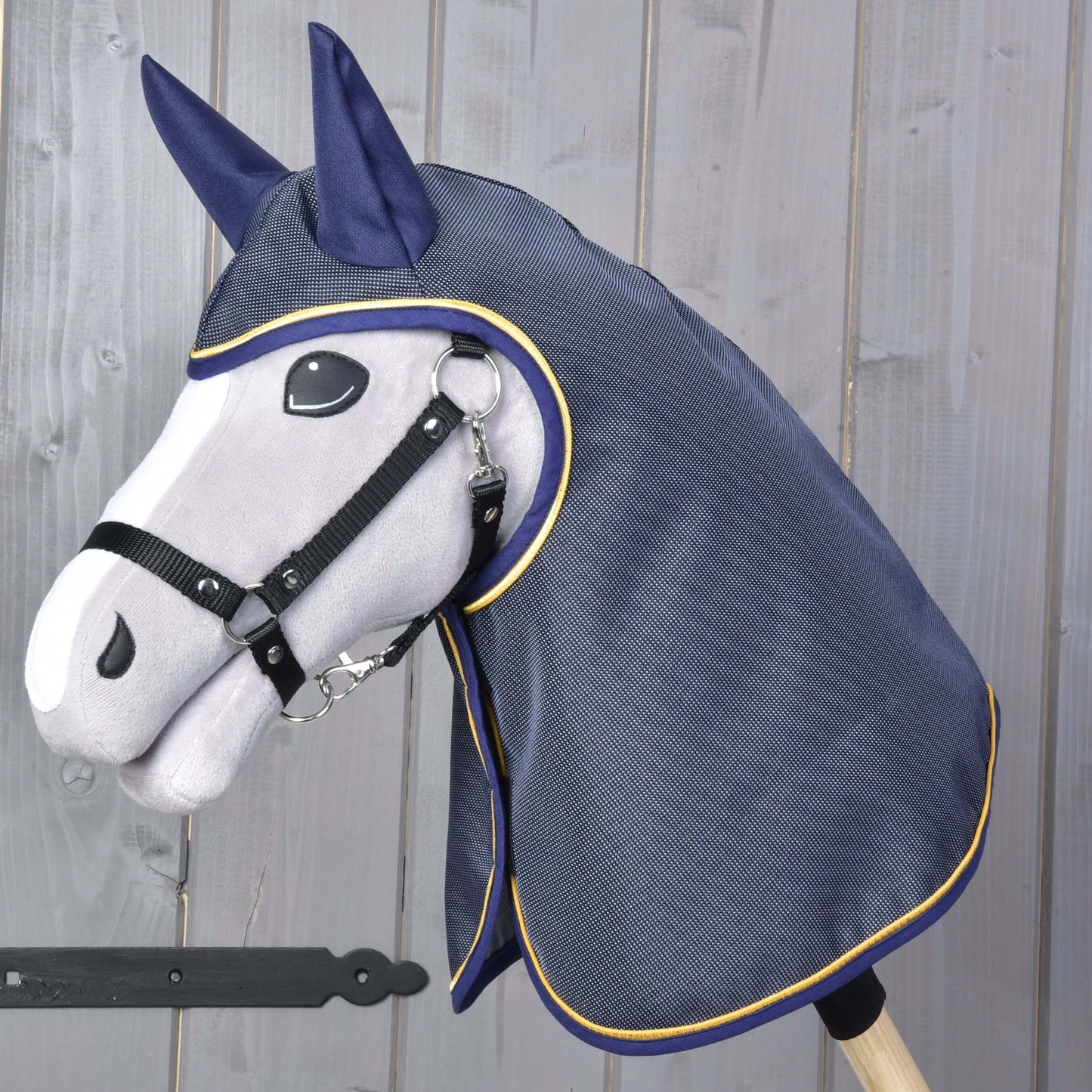 Pláštěnka Crone od Hobby Horse LarDen se šedou plyšovou hlavou Hobby Horse na tyči, černou uzdečkou a tmavě modrou pláštěnkou se žlutým lemováním, zobrazená na dřevěném pozadí.