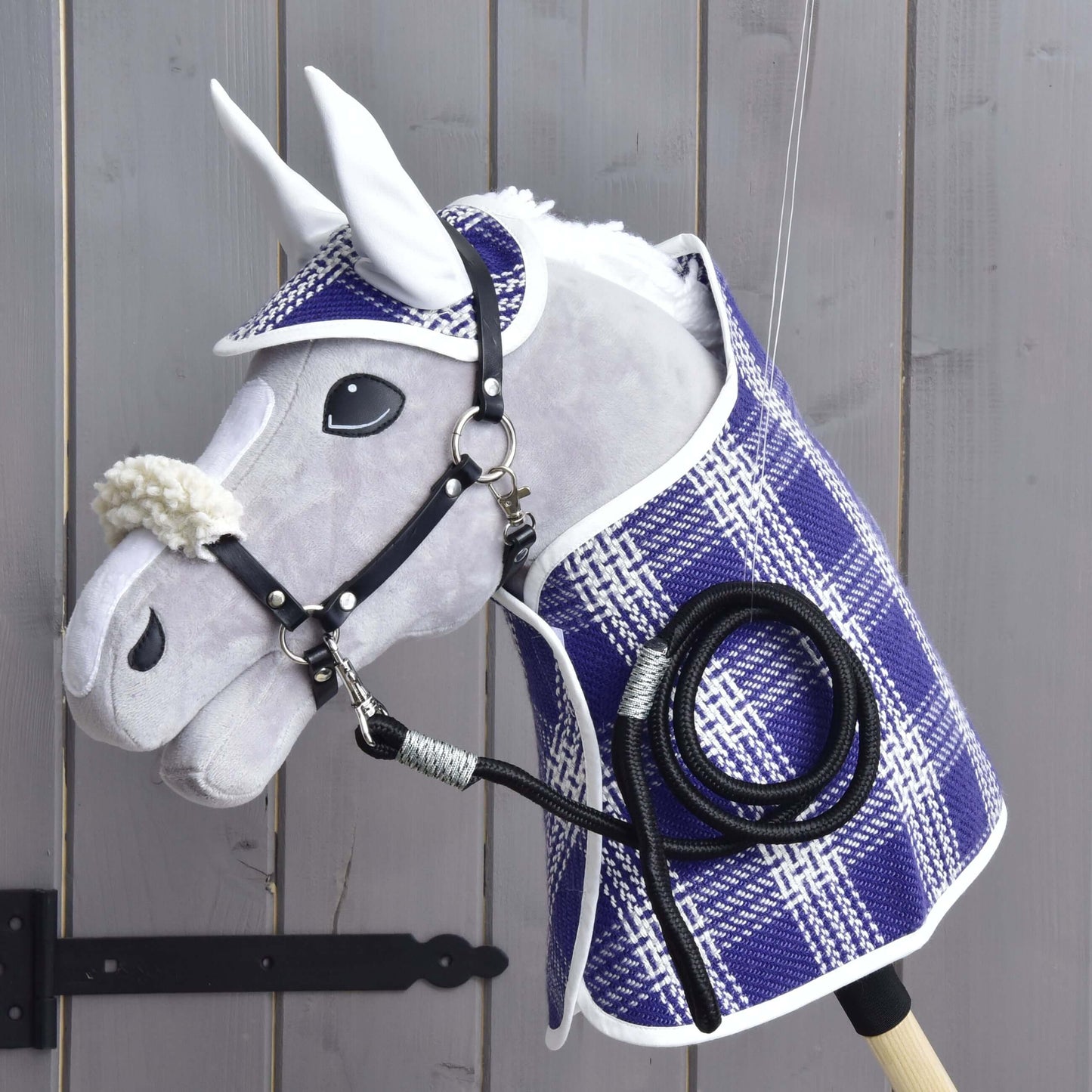 Set Cube (čabraka, deka, ohlávka s beránkem, vodítko) od Hobby Horse LarDen s dekou, čabrakou, vodítkem a ohlávkou s beránkem, vystavený na dřevěné stěně u černého pantu brány. Doplňky zdůrazňují kvalitu a kouzlo produktů Hobby Horse LarDen.