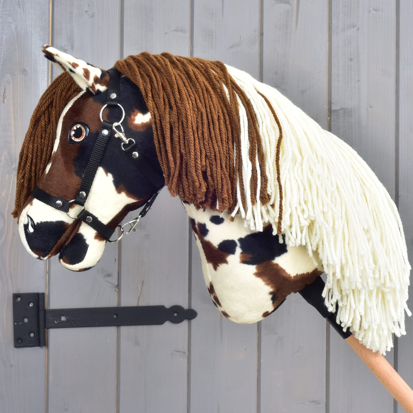 Hobby Horse Dakota s černou ohlávkou od Hobby Horse LarDen, ručně vyrobený model inspirovaný divokým duchem legendárních volně žijících koní Severní Ameriky. Má výrazné hnědé a bílé skvrny, měkkou hnědou přízovou hřívu, krémovou přízovou ofinu a je upevněný na dřevěné tyči; stojí před šedou dřevěnou stěnou jako pocta nezkrotnému mustangovi.