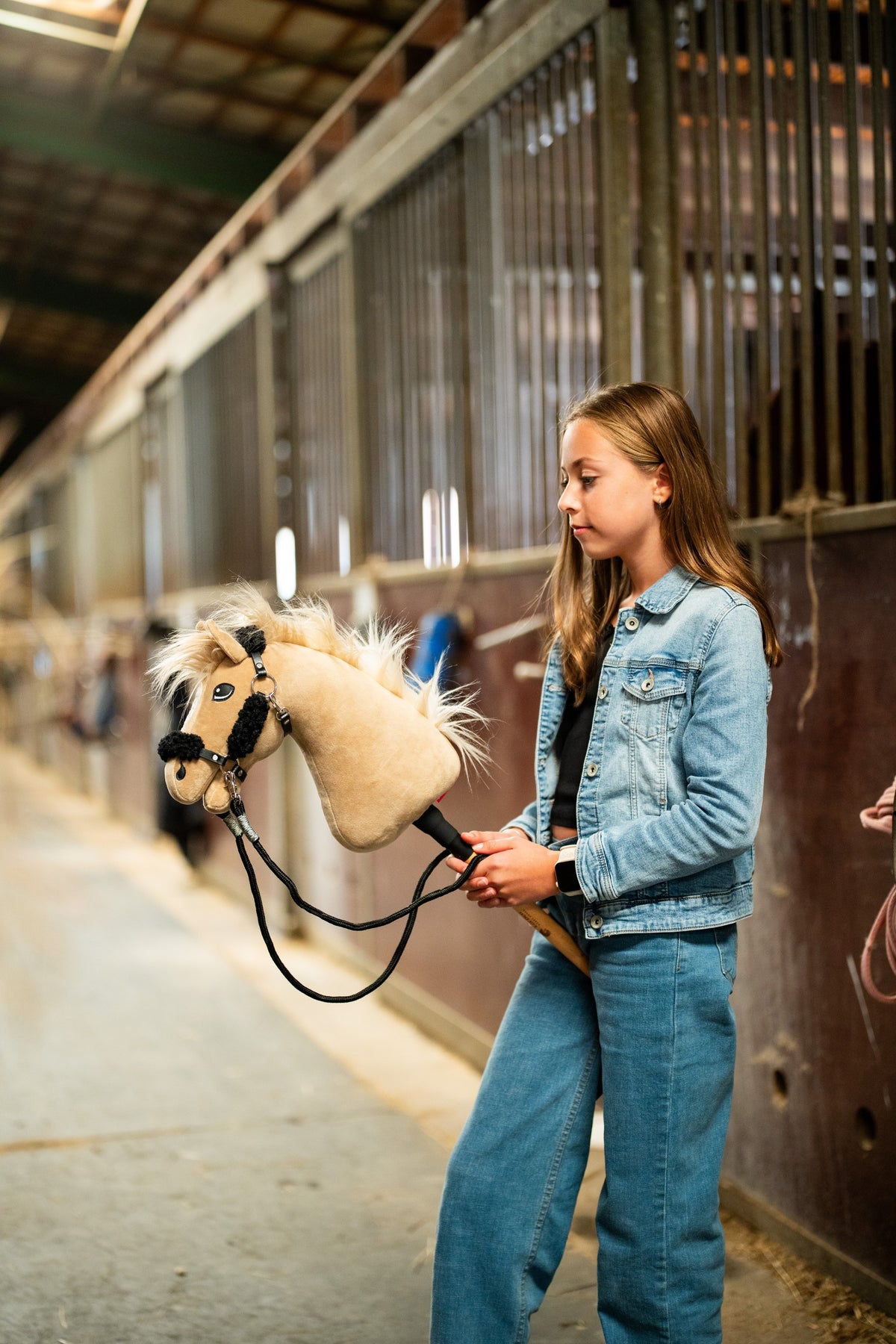 Dívka v džínové bundě vede uličkou stáje Hobby Horse Dancing Lady s černou ohlávkou od Hobby Horse LarDen, krémová plyšová hlava, světlá přízová hříva a ofina, s černou ohlávkou z koženky s kompletní černou kožešinou a černými otěžemi