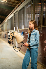 Dívka v džínové bundě vede uličkou stáje Hobby Horse Dancing Lady s černou ohlávkou od Hobby Horse LarDen, krémová plyšová hlava, světlá přízová hříva a ofina, s černou ohlávkou z koženky s kompletní černou kožešinou a černými otěžemi