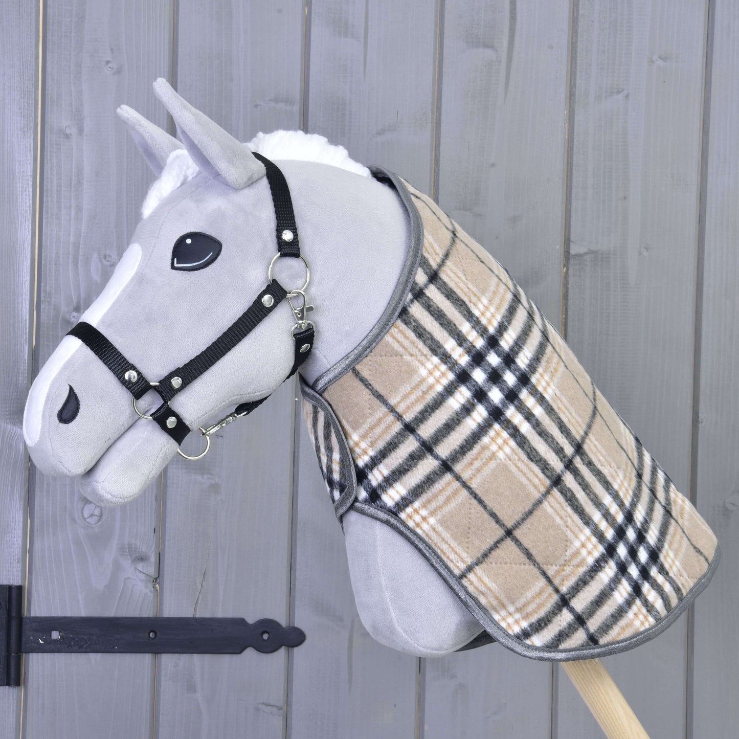 Hobby Horse LarDen „Deka Cremo England“ na šedém Hobby Horse s černou ohlávkou a béžovým kostkovaným kabátkem, doplněná čabrakou pro ochranu před hmyzem, nasazeném na dřevěné tyči před šedou dřevěnou stěnou.